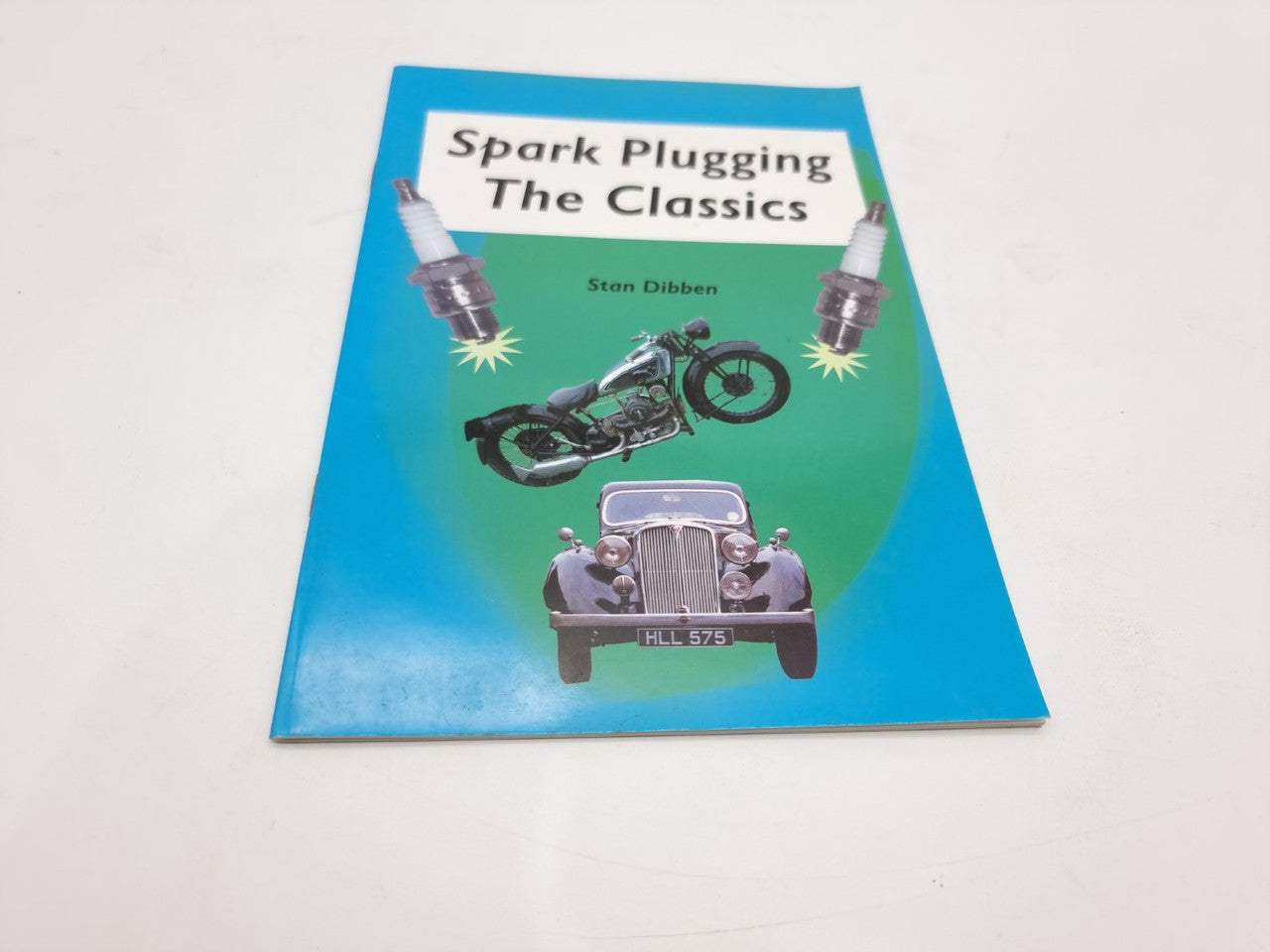 Paperback book cover with motorcycle and spark plugs, blue and green design, angled