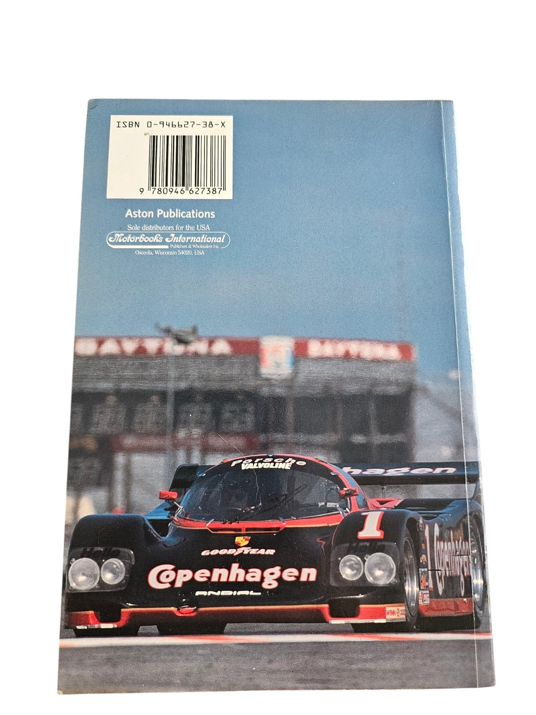 Back cover of Automoto Bookshop paperback featuring a black Copenhagen Porsche race car at Daytona speedway.